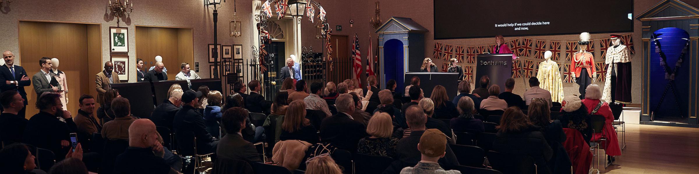 A crowd seated in a room with art and historical exhibits, listening to a auctioneer at the front