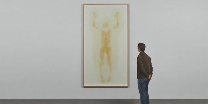 A man in a brown jacket looks at a large, framed artwork, showing a person with outstretched arms.