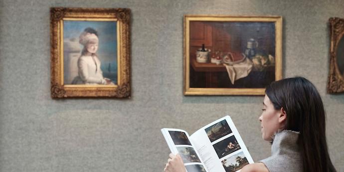 A woman reads an auction catalogue in front of two paintings with gold frames.